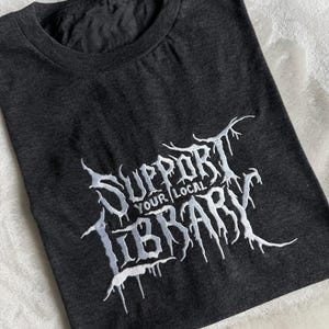 May include: A charcoal gray short-sleeve t-shirt featuring white embroidered text that reads SUPPORT YOUR LOCAL LIBRARY in a stylized, jagged, dripping font. The lettering has a textured appearance with sharp, pointed edges and elongated lines.