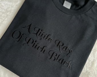 May include: Black t-shirt with the embroidered text "A Little Ray Of Pitch Black" in a decorative font. The shirt is folded and lies on a white, textured surface. The t-shirt is a solid black color.