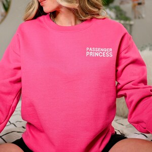 Op de afbeelding: Een roze sweatshirt met de tekst "PASSENGER PRINCESS" in witte letters op de voorkant.