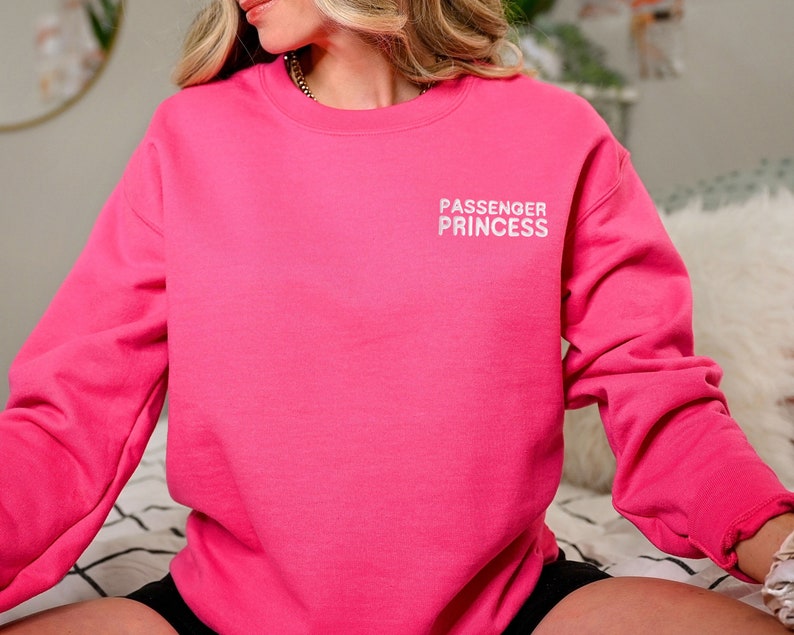 Op de afbeelding: Een roze sweatshirt met de tekst "PASSENGER PRINCESS" in witte letters op de voorkant.