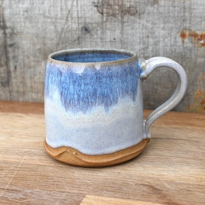 May include: A handmade ceramic mug with a blue and white glaze. The mug has a rounded body and a handle. The base of the mug is a light brown colour.