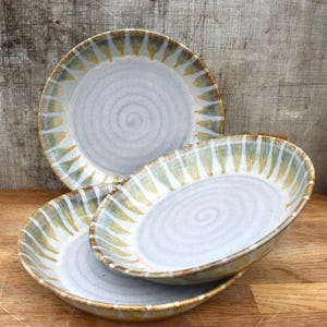 May include: Three handmade ceramic plates are stacked. The plates feature a light grey interior with a spiral design and a rim decorated with yellow and green brushstrokes. The plates are on a wooden surface.