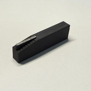 Pode incluir: Uma ferramenta de plástico preto com um inserto de metal. A ferramenta tem forma retangular e tem uma pequena superfície plana em uma extremidade.