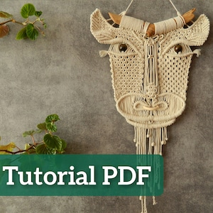 Op de afbeelding: Een beige macramé wandhanger in de vorm van een stierenkop. De stier heeft houten hoorns en donkerbruine kralen ogen. De tekst "Tutorial PDF" staat op een groene rechthoek. De achtergrond is een grijze muur.