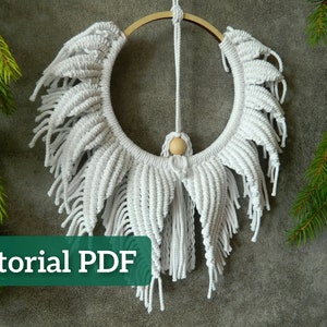 May include: White macrame wall hanging with a wooden ring and a wooden bead. The macrame is in the shape of a sunburst with long fringe. The text "Tutorial PDF" is at the bottom of the image.