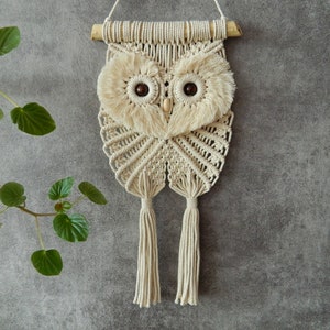 May include: A macrame owl wall hanging with wooden dowel and brown wooden bead eyes. The owl is made with natural colored string and has two long tassels.