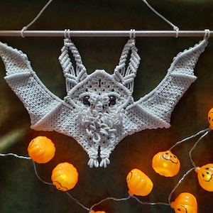 May include: A white macrame bat wall hanging with a wooden dowel at the top. The bat is hanging from a string and is surrounded by a string of orange pumpkin lights.