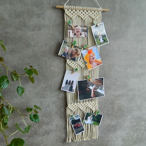 May include: Beige macrame photo display with a wooden dowel and cactus clips. It holds several photos, including images of people, a dog, a piano, and a red car. The macrame hangs against a grey wall.