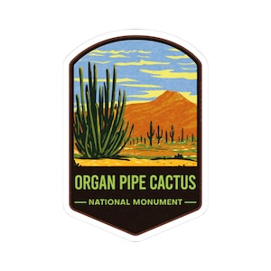 以下が含まれることがあります： オルガンパイプサボテン、山、青い空のある砂漠の風景を描いたステッカー。「ORGAN PIPE CACTUS - NATIONAL MONUMENT」の文字が緑と白で表示されています。