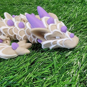 May include: A beige and white dragon figurine with purple accents. The dragon is lying on its belly with its head turned to the side. The dragon has a detailed, scaly body and a long, pointed tail.