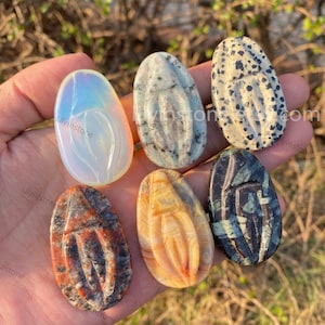 May include: A selection of six oval gemstones in different colours and patterns. The stones include a pearlescent white, speckled black and white, and others with brown, orange, and green hues. Each stone has a carved design.