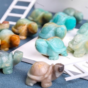 Puede incluir: Una colección de pequeñas figuritas de tortugas talladas en aventurina verde, marrón y blanca. Las tortugas están dispuestas sobre una superficie blanca con una escalera blanca en el fondo.