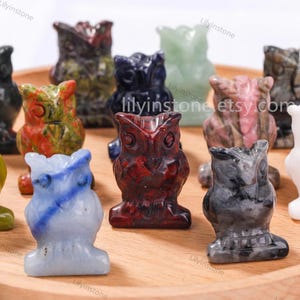 May include: A collection of carved owl figurines in various colors and stone textures. The owls are in shades of red, blue, green, and black. The figurines are displayed on a wooden tray.