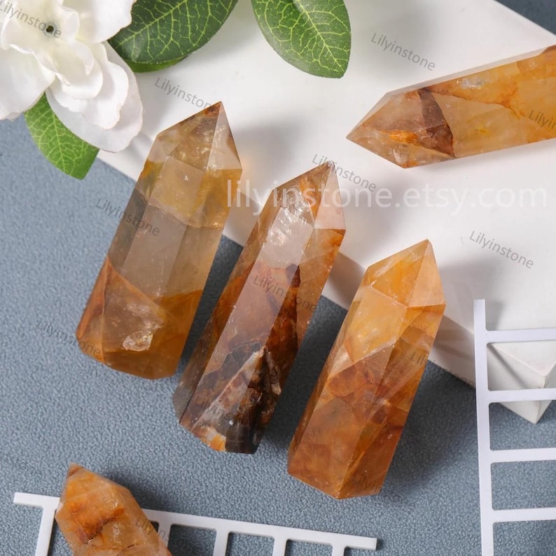 May include: Four golden citrine crystal points, each with a unique shape and pattern, are arranged on a white surface. The crystals are translucent and have a warm, golden hue. The image includes the text "lilyinstone.etsy.com".