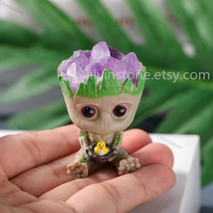 Peut inclure: Une petite figurine verte de Groot avec un cristal d'améthyste violet sur la tête. La figurine tient un petit oiseau jaune dans ses mains. La figurine est assise sur une main.