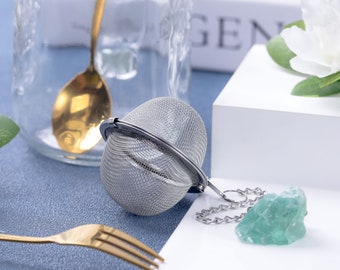 May include: A silver tea infuser ball with a chain, a green fluorite crystal, and a gold fork on a blue surface.