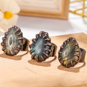 May include: Three rings with a copper finish and a green and gray gemstone set in a claw setting. The rings are on a wooden surface.