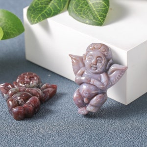May include: Two carved stone angel figurines. One is a light purple colour and the other is a reddish brown colour. Both are sitting with their wings spread out.