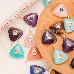 Puede incluir: Una colección de colgantes triangulares de piedras preciosas en varios colores, incluyendo púrpura, rosa y turquesa. Cada colgante presenta una argolla dorada y una gema circular central. Los colgantes se exhiben sobre una superficie de madera.