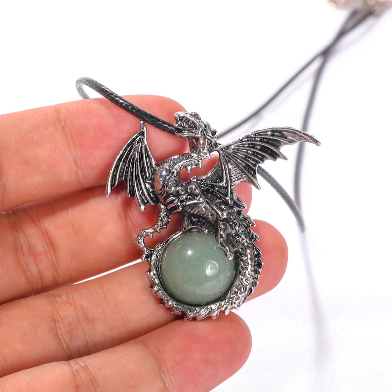 May include: A silver dragon pendant with a green gemstone. The dragon is curled around the stone, and the pendant is attached to a black cord.