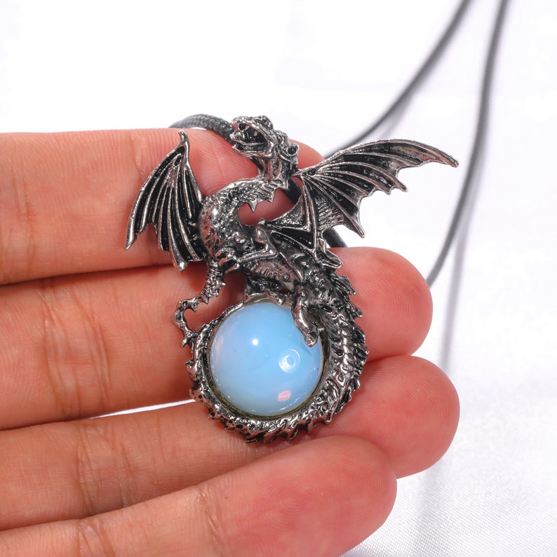 May include: A silver dragon pendant with a blue opal stone. The dragon is curled around the stone, with its wings spread out. The pendant is on a black cord.