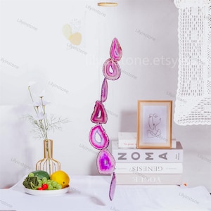 May include: A pink agate crystal wind chime with six hanging pieces. The chime is suspended from a wooden ring and hangs from a white ceiling.