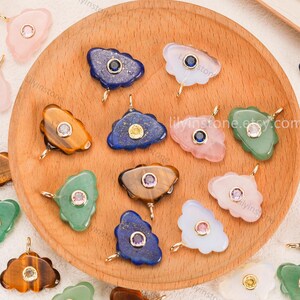 May include: A collection of cloud-shaped pendants crafted from various gemstones including lapis lazuli, tiger's eye, rose quartz, and green aventurine. Each pendant features a central round faceted gemstone set in a gold-toned metal bezel.