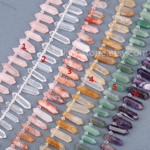 May include: Six strands of faceted gemstone beads in various colors, including clear, pink, red, yellow, green, and purple. The beads are arranged in rows and are all in the shape of a long, pointed teardrop. The beads are strung on a clear string.