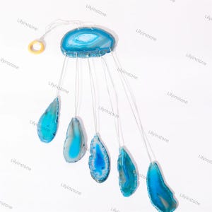May include: A blue agate wind chime with six pieces of agate hanging from a circular piece of agate. The agate pieces are all different shapes and sizes. The wind chime is hanging from a white string.