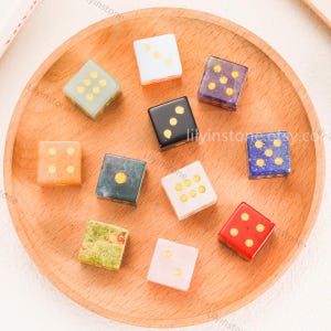 May include: A collection of ten square dice in various colors and materials, including white, black, red, blue, green, and marbled patterns, each featuring gold-colored circular pips on the top faces, arranged on a round wooden tray.