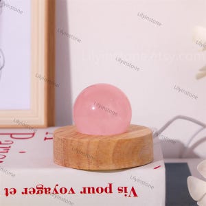 May include: A pink rose quartz sphere on a wooden stand. The sphere is smooth and polished. The stand is made of natural wood.