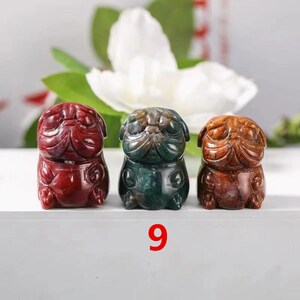 May include: Three small carved stone figurines of pugs in red, green, and brown. The figurines are sitting on a white surface with the number 9 printed below them.