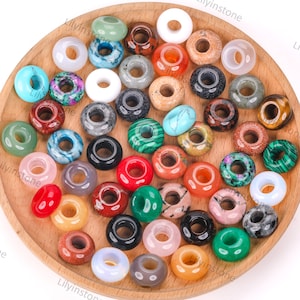 Op de afbeelding: Een verzameling donutvormige edelsteen kralen in diverse kleuren, waaronder groen, rood, blauw en oranje, gepresenteerd op een houten dienblad. De kralen hebben een gat in het midden en variëren in textuur en patroon.