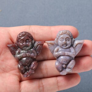 May include: Two carved cherub figurines made of red and purple agate. The cherubs are sitting with their wings spread out and their hands clasped in front of them.