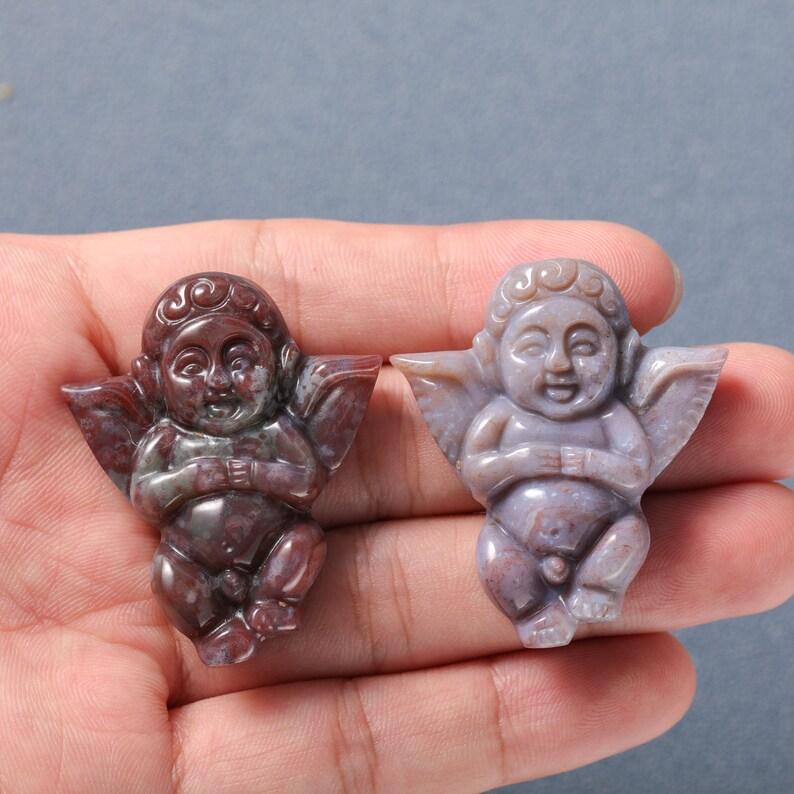 May include: Two carved cherub figurines made of red and purple agate. The cherubs are sitting with their wings spread out and their hands clasped in front of them.