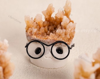 Puede incluir: Un objeto decorativo artesanal y caprichoso. Presenta una base blanca con una cara sonriente y gafas de montura negra. La parte superior está llena de un grupo de cristales de color naranja claro, creando una exhibición artística única.