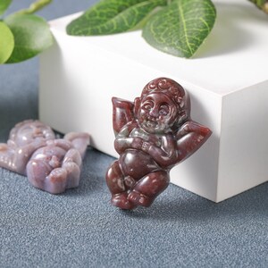 May include: Two carved stone figurines of a cherub with wings. The figurine on the left is a light purple colour and the figurine on the right is a reddish brown colour.