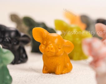 May include: A collection of small, carved pig figurines in various colors, including orange, green, black, and pink. The focus is on a detailed orange pig figurine with large ears and a smiling face. The background is blurred, with other figurines visible.
