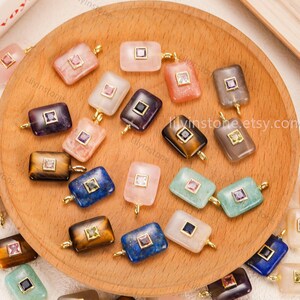 May include: A collection of rectangular gemstone pendants in various colors, including pink, blue, green, and brown, each with a square inset and gold-colored bail. The pendants are displayed on a wooden plate.