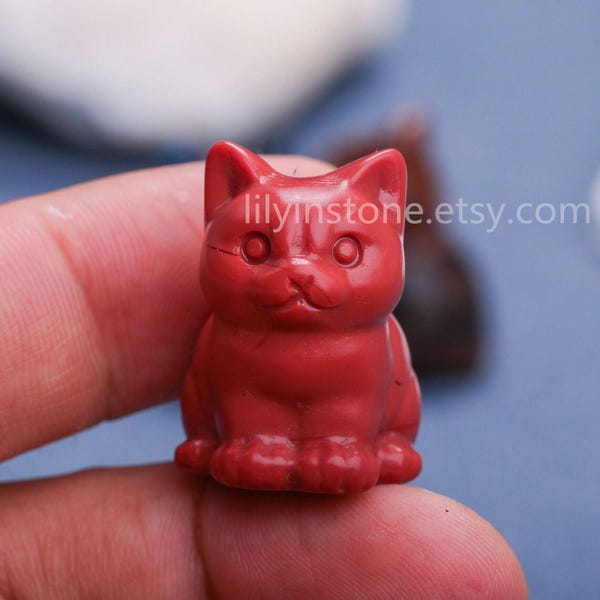 Op de afbeelding: Een klein, rood, gesneden kattenbeeldje. De kat zit met zijn poten onder zijn lichaam. Het beeldje is gemaakt van een glad, gepolijst materiaal.