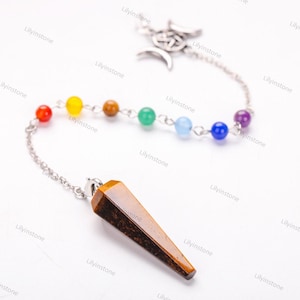 Op de afbeelding: Een gouden slinger met een punt van tijgeroogkristal en een ketting met zeven chakra-parels. De parels zijn rood, oranje, geel, groen, blauw, indigo en violet.