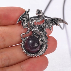 May include: Silver dragon pendant with a purple gemstone in the center. The dragon is holding the gemstone in its claws. The pendant is on a silver chain.