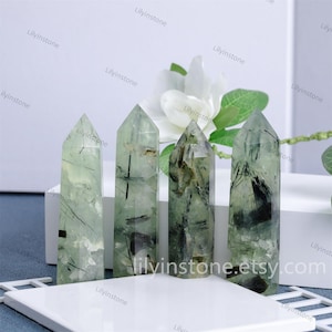 Puede incluir: Cuatro puntas de cristal verde y negro, también conocidas como cuarzo fantasma verde, están dispuestas sobre una superficie blanca. Los cristales son translúcidos y tienen una textura natural y rugosa.