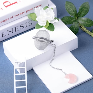 May include: A silver tea infuser ball with a chain and a pink crescent moon-shaped crystal charm. The infuser is on a white surface with a white ladder and a book with the word "GENESIS" on the cover.