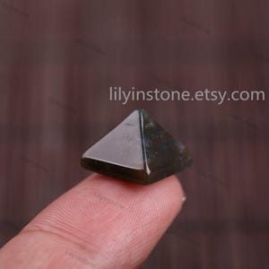 Peut inclure: Un petit cristal en forme de pyramide vert fonc&eacute;, avec une surface lisse et polie. Le cristal est tenu par un doigt, et l'arri&egrave;re-plan est de couleur brune floue.