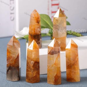 May include: Six orange and brown crystal points, also known as citrine, on a white surface. The points are all different sizes and shapes, but they are all polished and have a smooth finish.