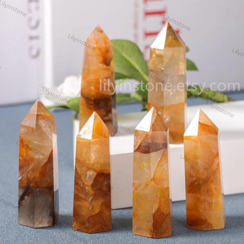 May include: Six orange and brown crystal points, also known as citrine, on a white surface. The points are all different sizes and shapes, but they are all polished and have a smooth finish.