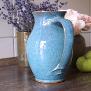 Könnte beinhalten: Eine türkisfarbene Keramikkrug mit gebogenem Griff, auf einer Holzoberfläche. Der Krug steht neben einer Vase mit Lavendel, einer Vase mit rosa Blumen und zwei grünen Äpfeln. Der Krug hat eine strukturierte Oberfläche.