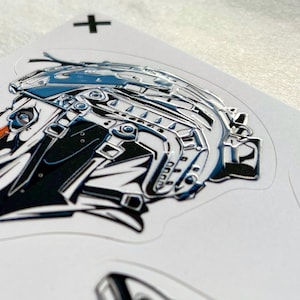 Puede incluir: Una pegatina brillante con una ilustraci&oacute;n estilizada de una cabeza de p&aacute;jaro con un casco t&aacute;ctico en tonos azul, negro y blanco con un detalle de pico naranja. Un peque&ntilde;o signo m&aacute;s negro est&aacute; impreso en el soporte blanco cerca del borde.