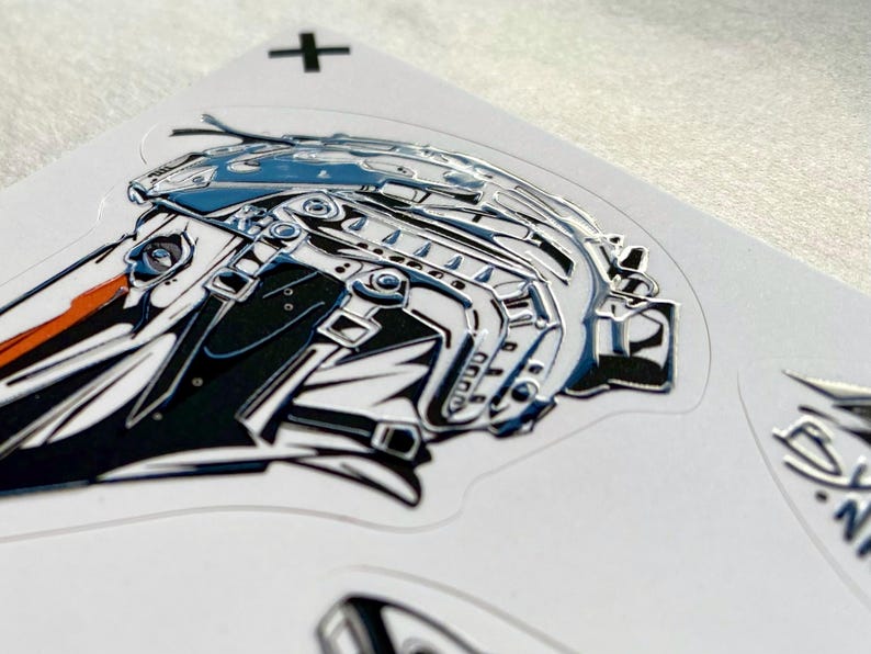 Puede incluir: Una pegatina brillante con una ilustraci&oacute;n estilizada de una cabeza de p&aacute;jaro con un casco t&aacute;ctico en tonos azul, negro y blanco con un detalle de pico naranja. Un peque&ntilde;o signo m&aacute;s negro est&aacute; impreso en el soporte blanco cerca del borde.